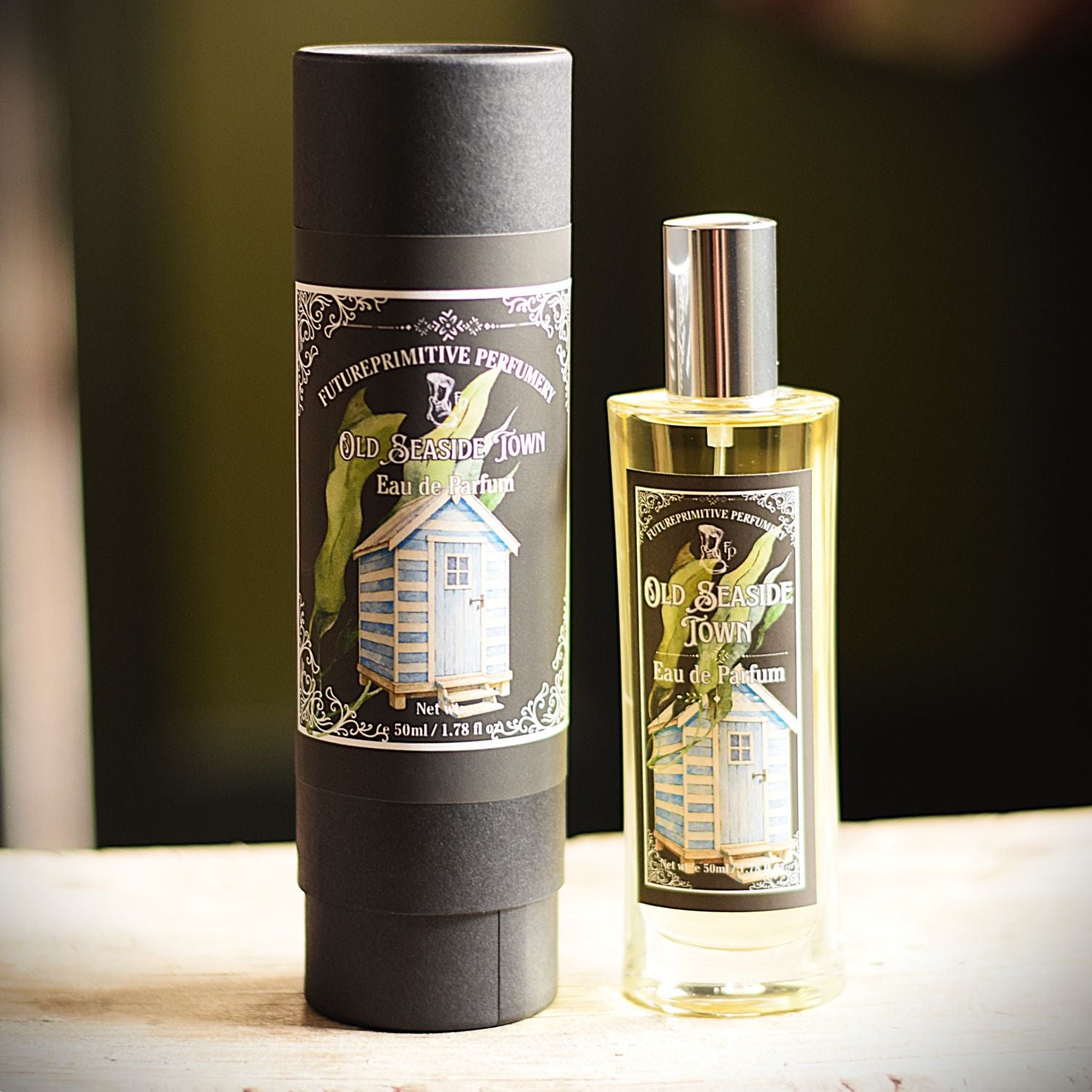 A bottle of perfume and its cylindrical packaging box, both labeled 'FUTUREPRIMITIVE PERFUMES Old seaside town Eau de Parfum', standing on a light wooden surface with a dark, blurred background.