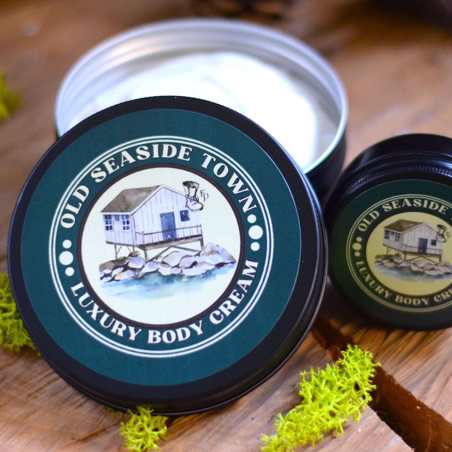 Two containers of Old Seaside Town Luxury Body Cream on a wooden surface with greenery.