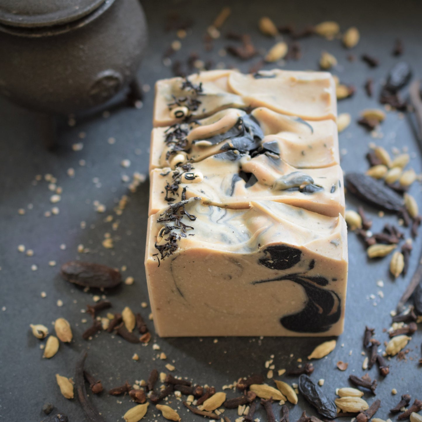 A rectangular bar of handmade soap with a light beige base and dark charcoal swirls, decorated with dried herbs, set on a dark slate surface with scattered spices and a dark ceramic pot in the background.