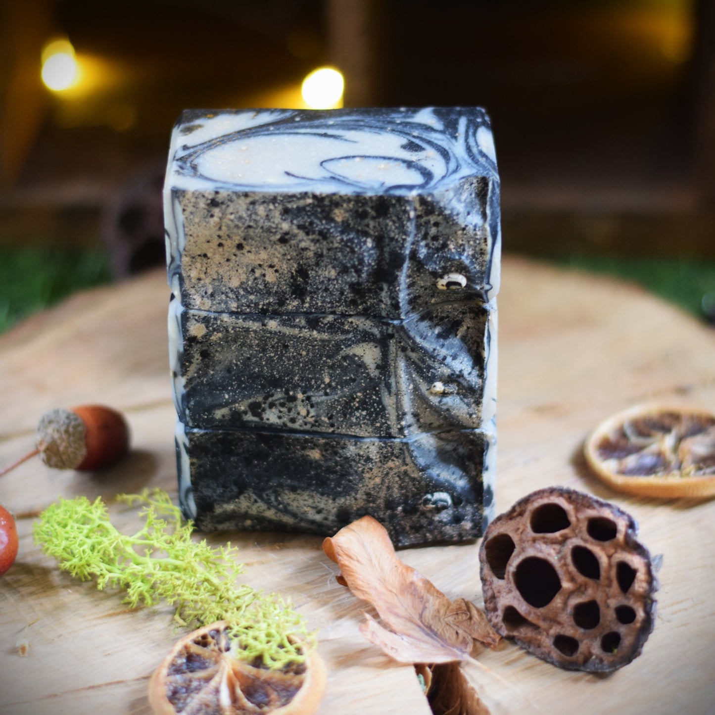 A rectangular bar of handmade soap with a black and white marble pattern, displayed on a rustic wooden surface surrounded by dried botanicals and moss.