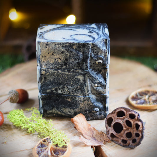 A rectangular bar of handmade soap with a black and white marble pattern, displayed on a rustic wooden surface surrounded by dried botanicals and moss.
