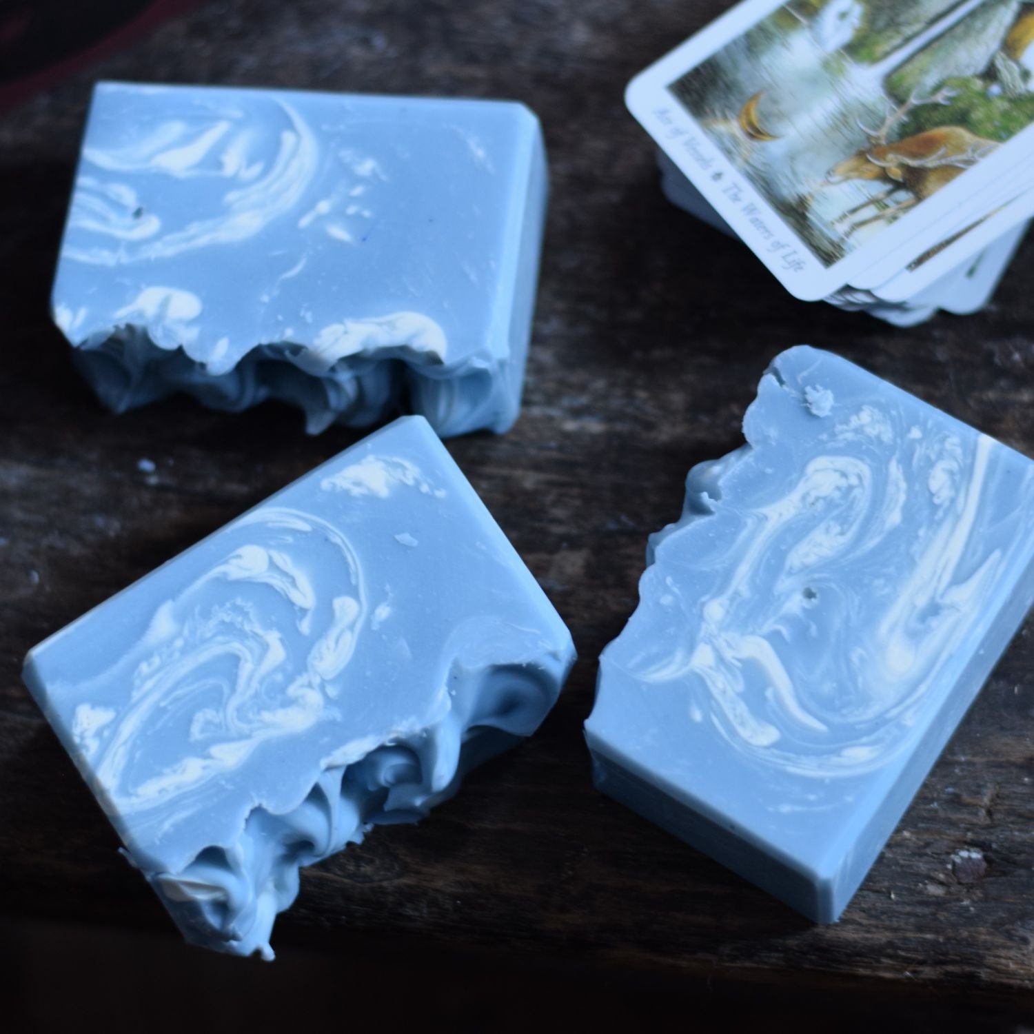 Three blue marbled soap bars on a dark wooden surface with a tarot card in the background.