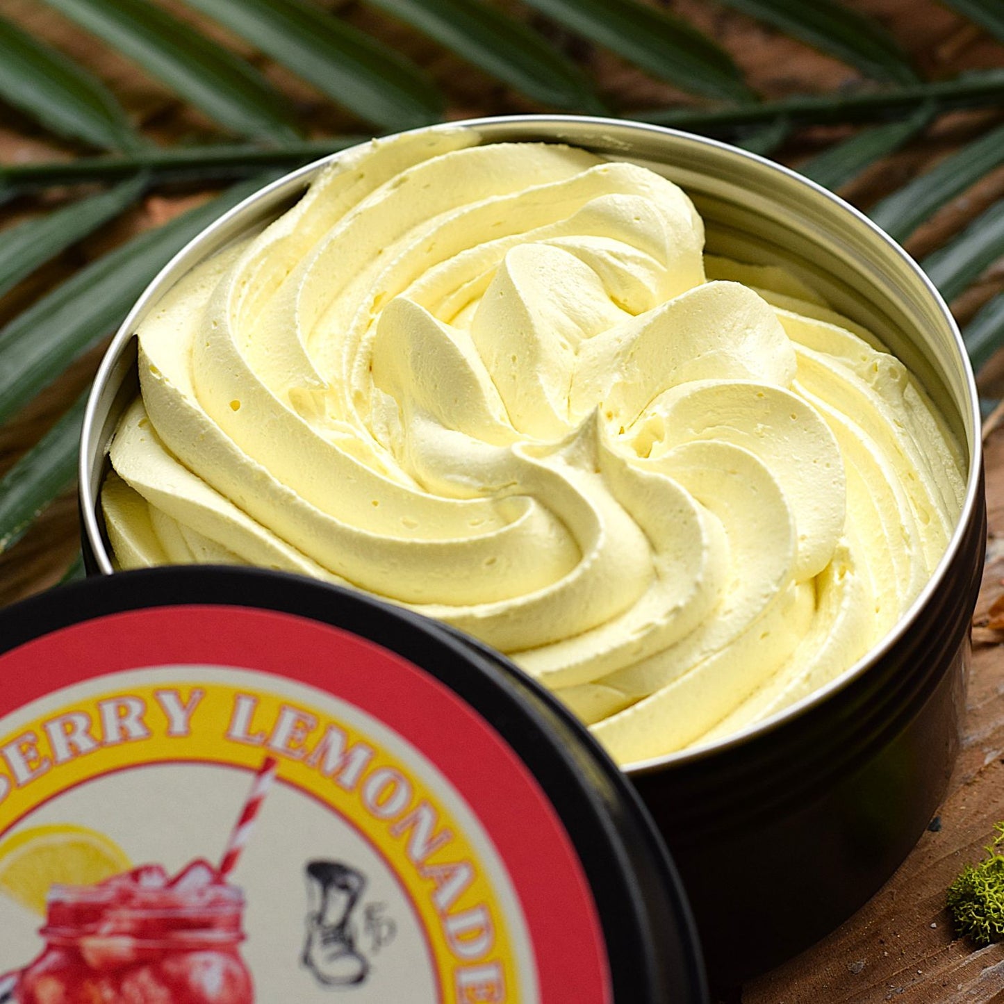 A close-up of a whipped cream soap wirled in a round metal tin, with the lid partly visible in the foreground. The lid label reads Raspberry Lemonade.