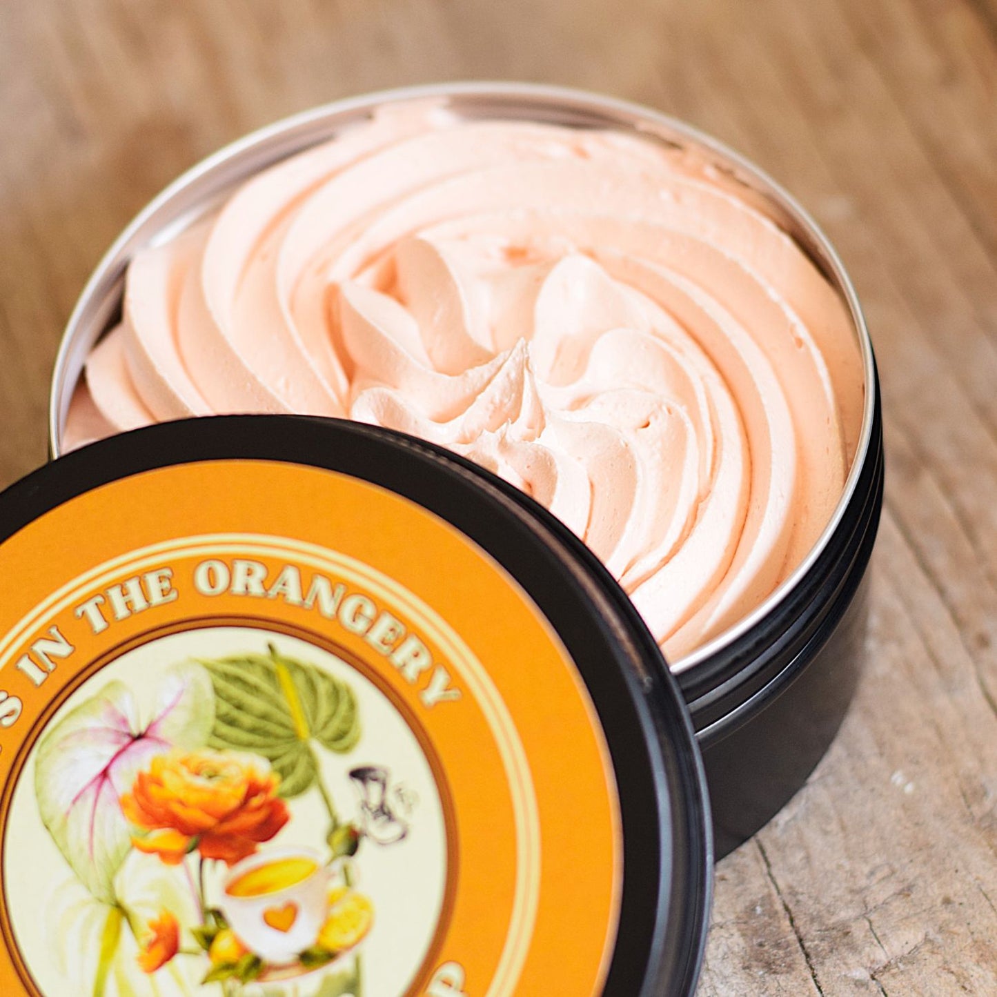 An open round metal tin filled with pale peach whipped cream or balm, shown on a wooden surface with the branded lid placed in front. The lid label reads "SHE'S IN THE ORANGERY" and features orange, pink, green, and cream floral artwork.