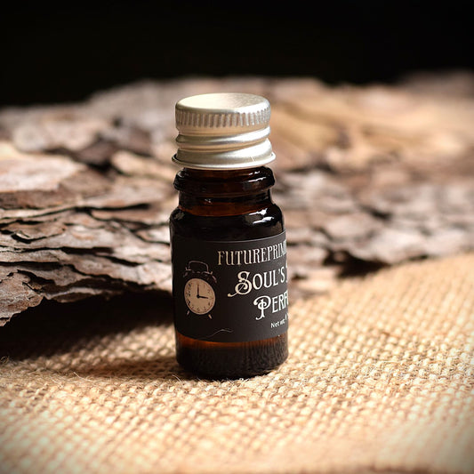 A small amber glass bottle with a silver cap and a black label reading 'FUTUREPRIMITIVE Perfume' sits on a textured burlap surface with rustic wood in the background.