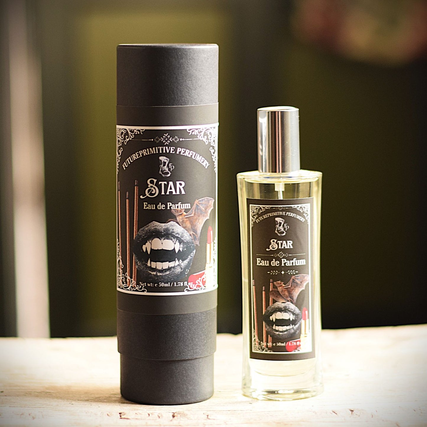 A bottle of Star Eau de Parfum with a silver cap stands next to its tall black cylindrical packaging tube on a light wooden surface, with a blurred dark green background.