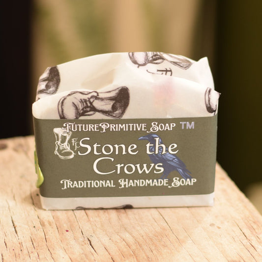 A wrapped bar of soap on a rustic wooden surface, with a dark olive-green label reading 'Ft. Stone the Crows Traditional Handmade Soap' and white paper wrapping printed with black crow illustrations.