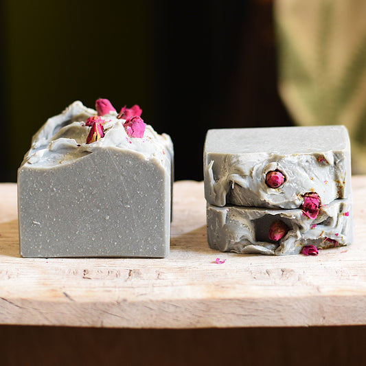 Two rectangular bars of handmade soap in a matte gray color, one standing upright and the other stacked horizontally, both decorated with dried pink rose petals, displayed on a rustic wooden surface.
