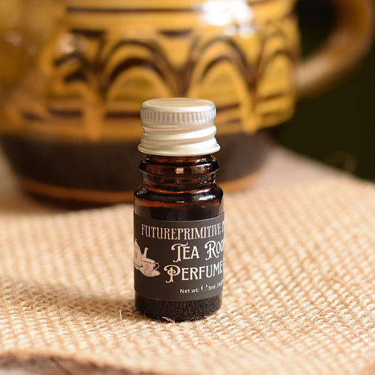 A small amber glass bottle of Future Primitive Tea Rooms Perfume with a silver cap, set on a burlap surface with a blurred ceramic teapot in the background.