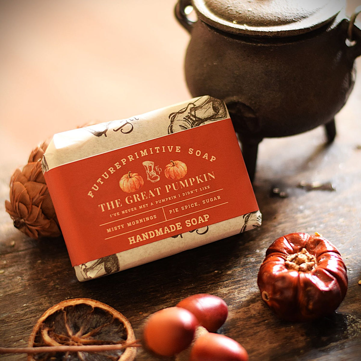 A bar of handmade soap wrapped in a terracotta orange label and cream paper with pumpkin illustrations, styled on a rustic wooden table with acorns, a dried orange slice, and a dark cast iron pot in the background.