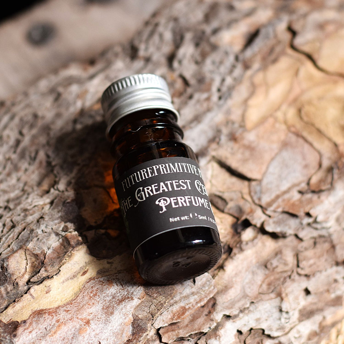 A small amber glass bottle of Future Primitive 'The Greatest Cedar of All Perfume' with a silver cap and black label, resting on textured tree bark.