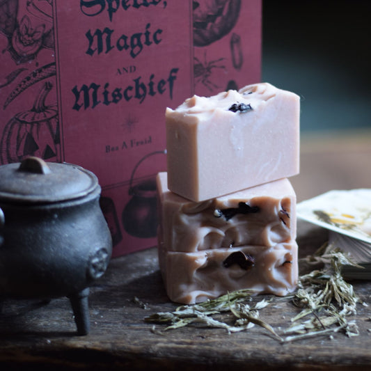 Two stacked soap bars with dried herbs on a wooden surface, with a book titled 'Spells, Magic and Mischief' in the background.