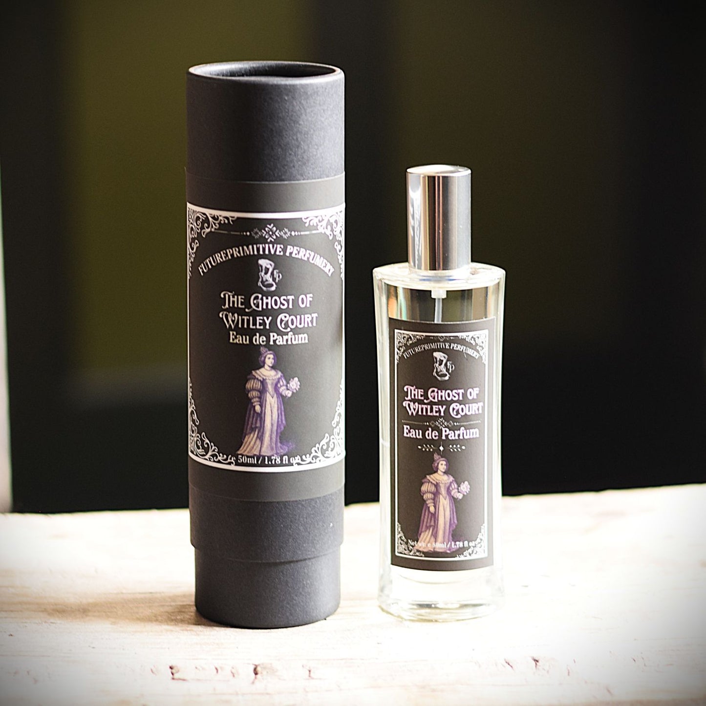 A bottle of Eau de Parfum and its matching matte black cylindrical box, both featuring a label for 'The Ghost of Witley Court' by 'FuturePrimitive Perfumery', displayed on a light wooden surface against a dark background.