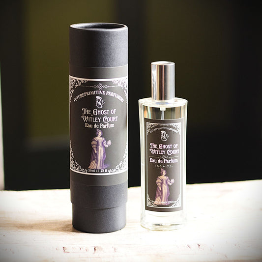 A bottle of Eau de Parfum and its matching matte black cylindrical box, both featuring a label for 'The Ghost of Witley Court' by 'FuturePrimitive Perfumery', displayed on a light wooden surface against a dark background.