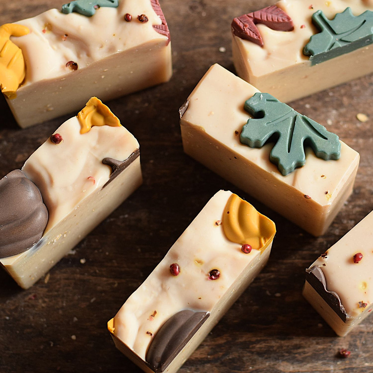 Several rectangular bars of artisanal soap arranged on a dark wooden surface. The soaps are light cream with decorative autumn-themed toppings including teal and yellow leaf shapes, small red berry-like accents, and dark brown swirls.