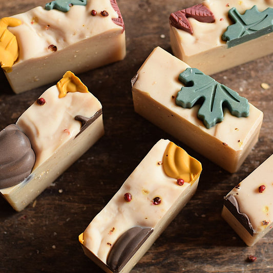 Several rectangular bars of artisanal soap arranged on a dark wooden surface. The soaps are light cream with decorative autumn-themed toppings including teal and yellow leaf shapes, small red berry-like accents, and dark brown swirls.