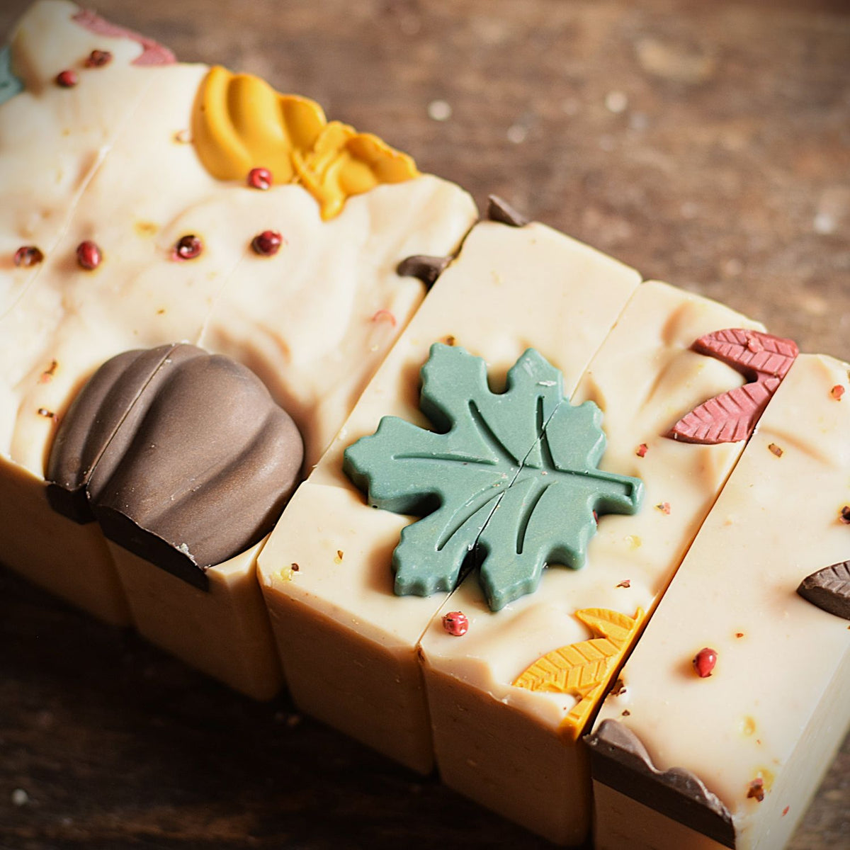 Close-up of a decorative bar of soap with a warm cream base, autumn-themed embossed details including a green maple leaf, yellow leaf, pink leaf, brown acorn, and small red dots, photographed on a rustic wooden surface.
