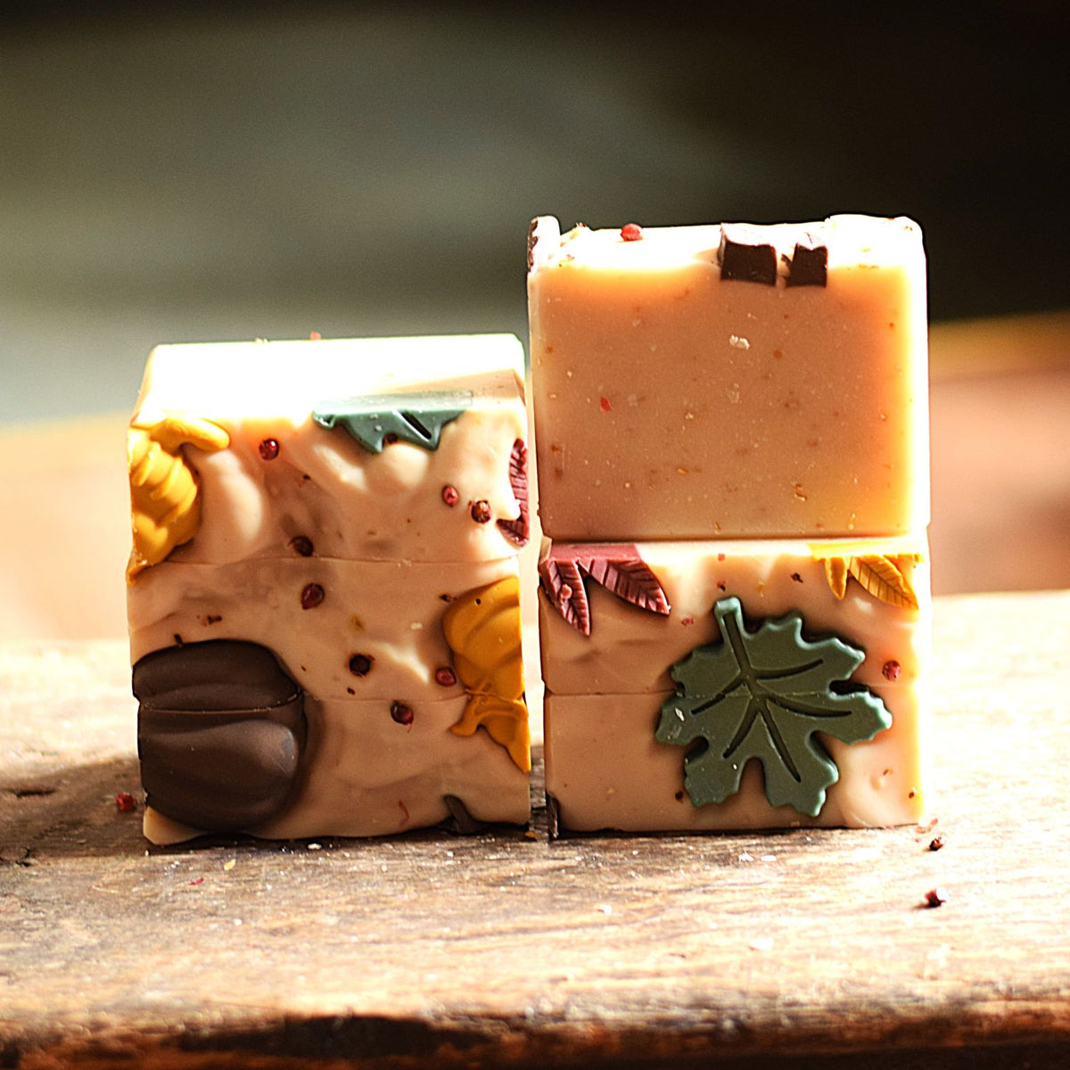 Two decorative bars of handmade soap standing upright on a rustic wooden surface. One bar is a light cream color with embedded yellow and green autumn leaves and a brown acorn detail, while the other is a warm peach-beige color with a green leaf and small dark accents.