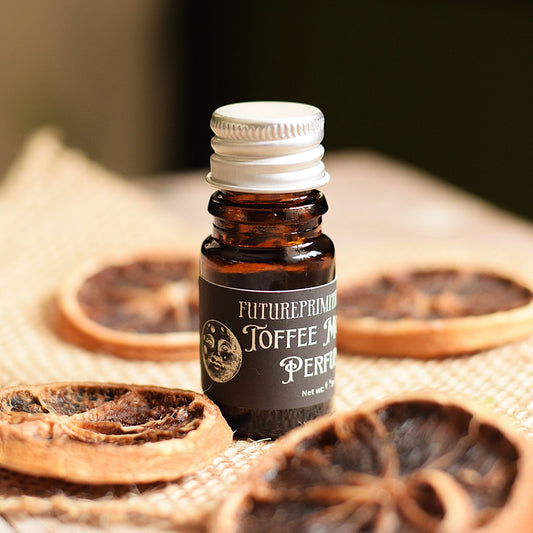 A small amber glass bottle with a white cap and a dark label reading 'FUTUREPRIMITIVE TOFFEE MOON FACE PERFUME OIL' stands on a burlap surface with dried coffee slices around it.