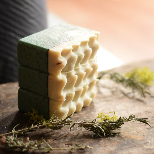 Stack of green and beige artisan soap bars on a wooden surface with herbs.