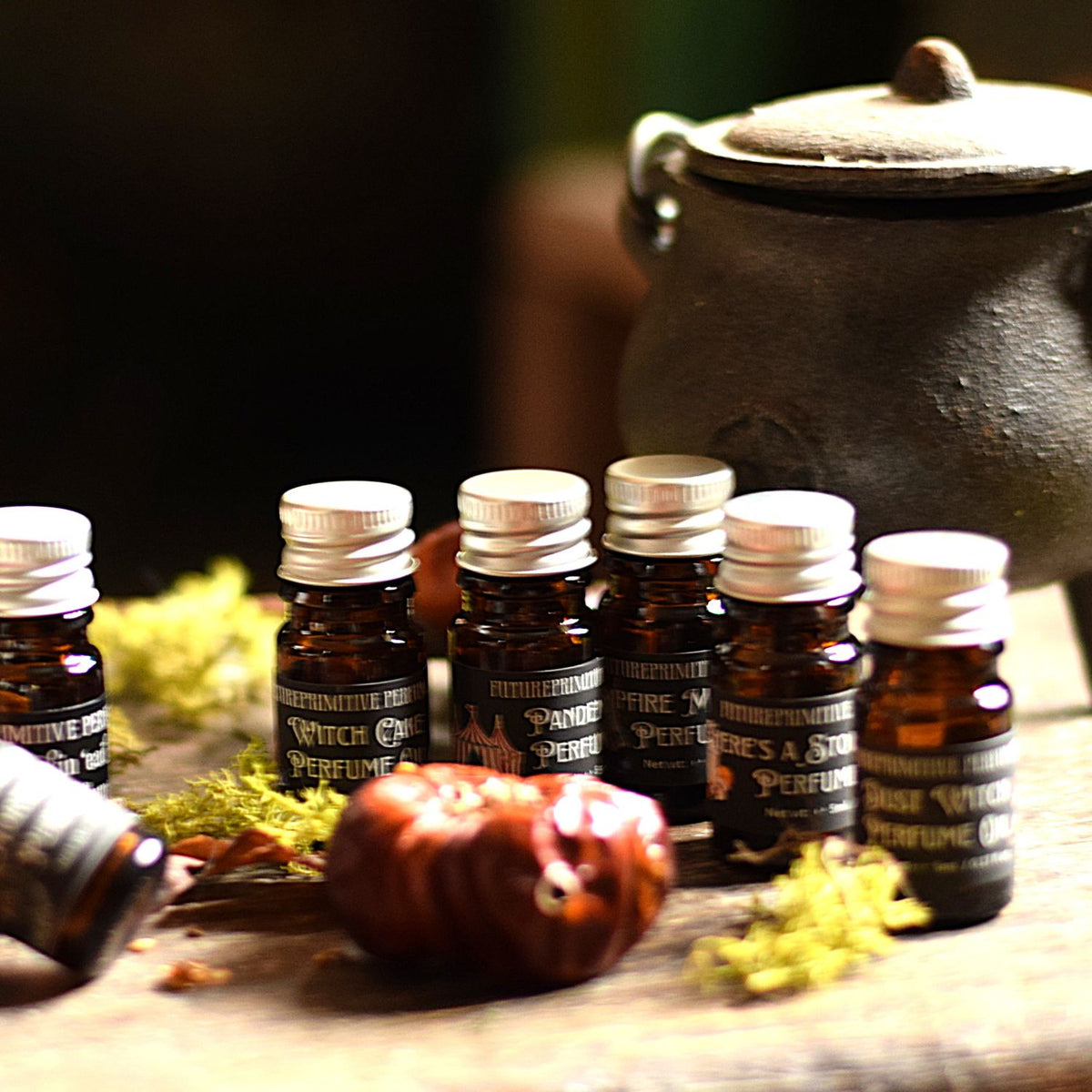 Six amber glass perfume bottles with silver caps and black labels arranged on a wooden surface, with a dark cauldron in the background and dried botanical props around them.