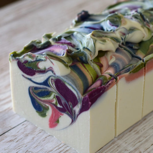 A close-up of a rectangular bar of handmade soap with a marbled swirl pattern of purple, green, blue, pink, and cream on a light wood background.