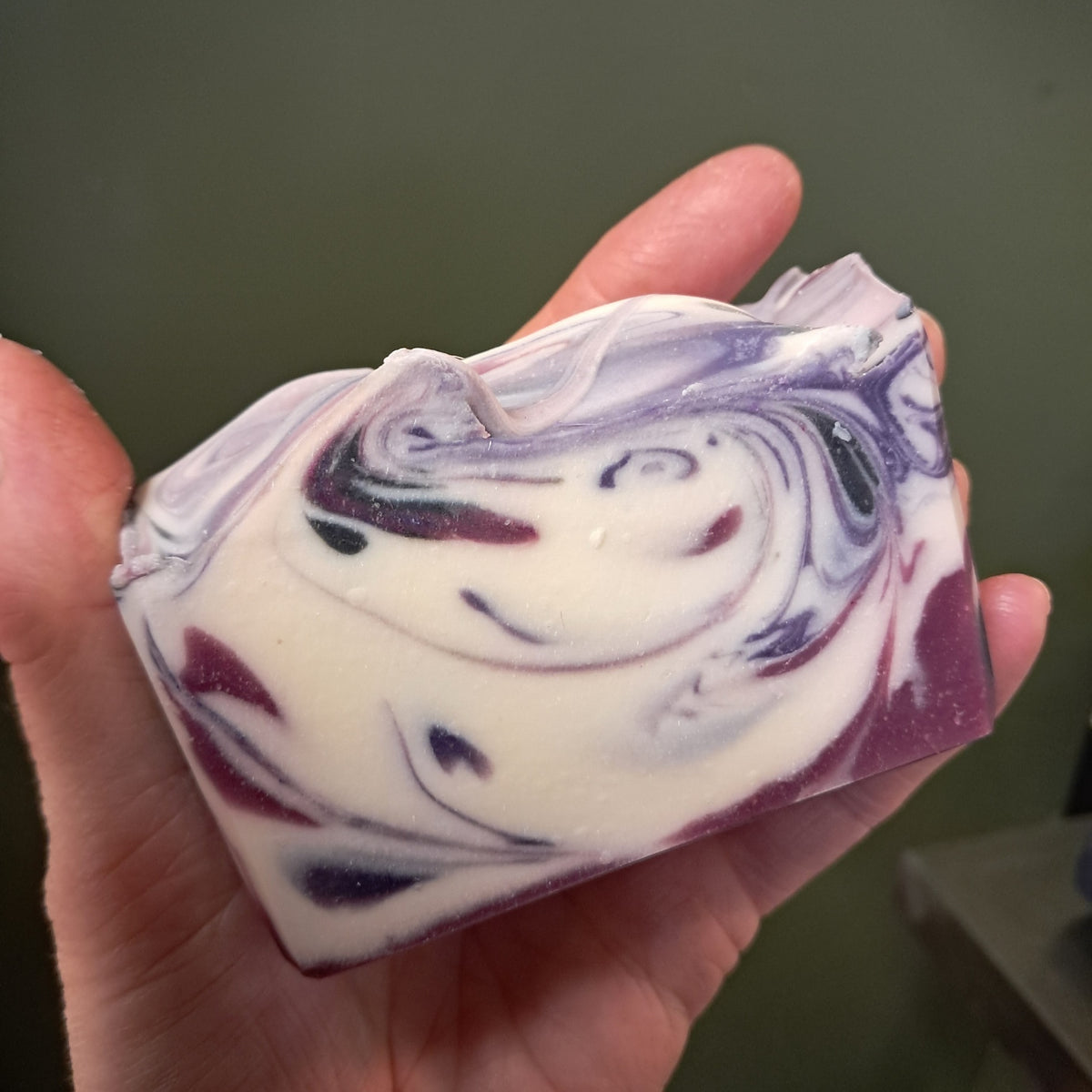 A hand holding a rectangular bar of handmade soap with a marbled swirl pattern of purple, white, and dark red, set against a dark olive green background.