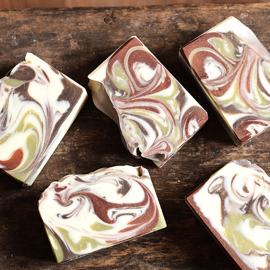 Five artisanal marbled soap bars with swirls of cream, brown, olive green, and white, arranged on a rustic dark wooden surface.